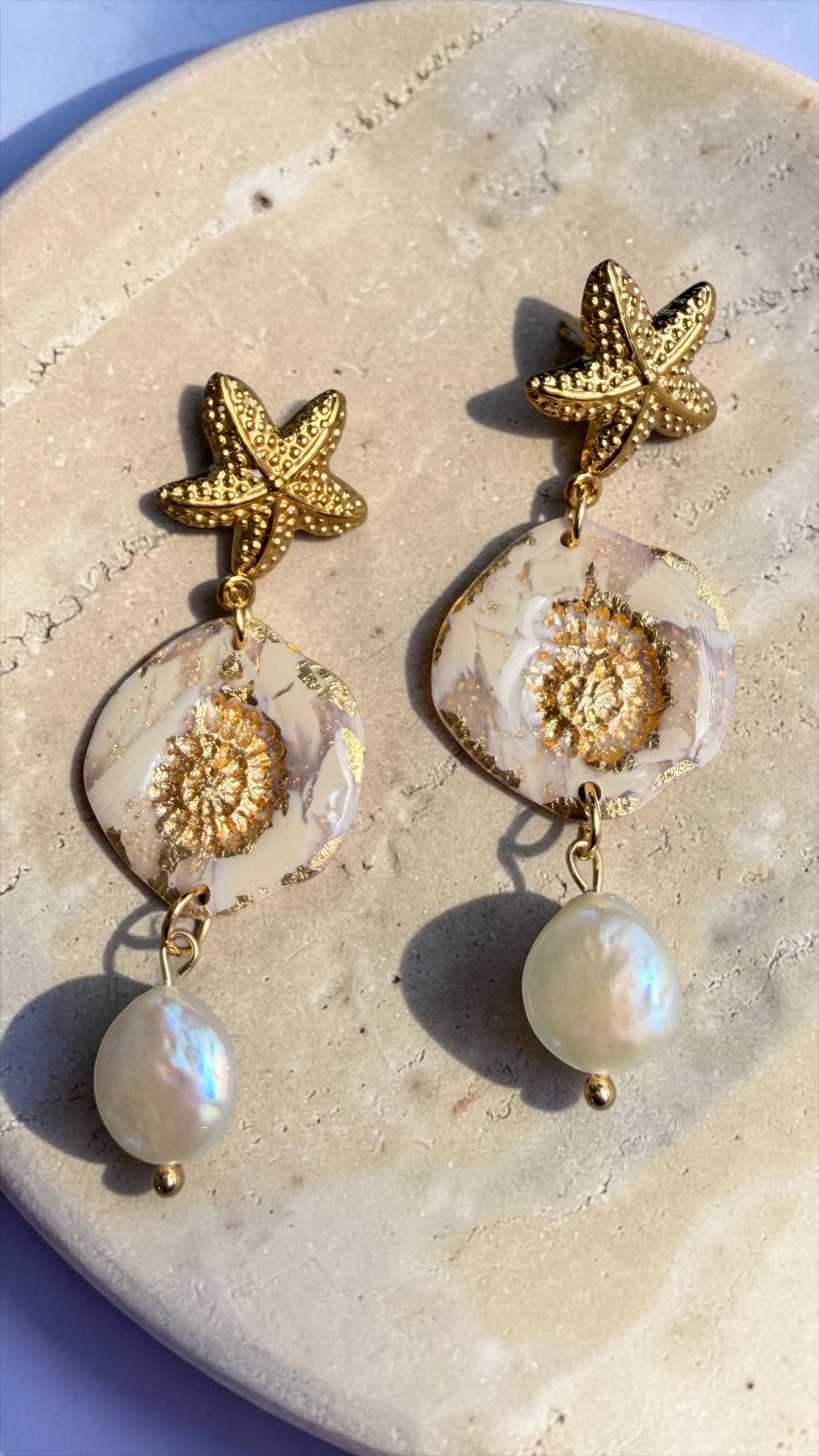 Ohrringe Nerea Pearl mit goldenem Seestern, marmorierter Polymer-Anhänger und Süsswasserperle, fotografiert auf Travertinplatte