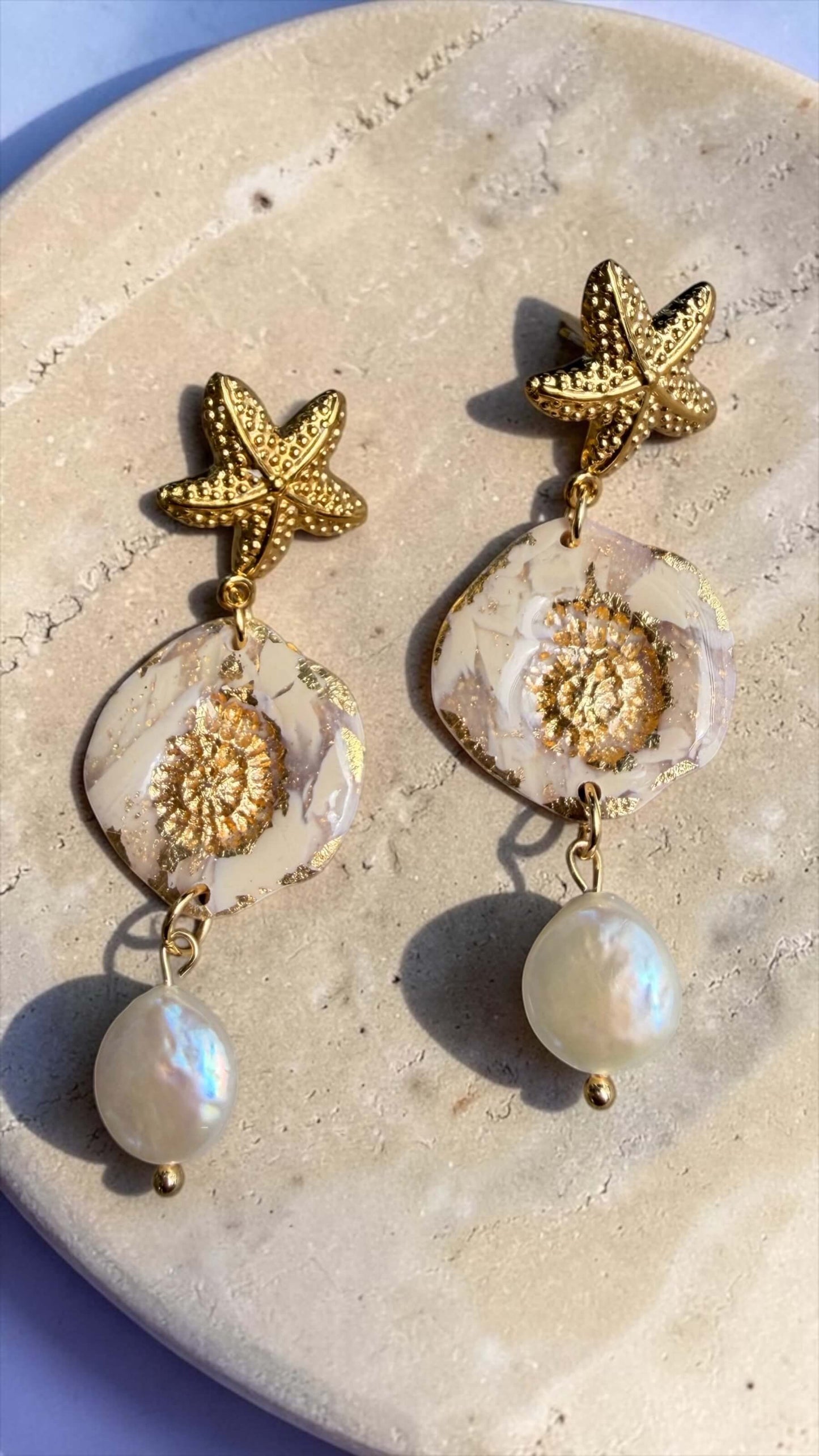 Ohrringe Nerea Pearl mit goldenem Seestern, marmorierter Polymer-Anhänger und Süsswasserperle, fotografiert auf Travertinplatte