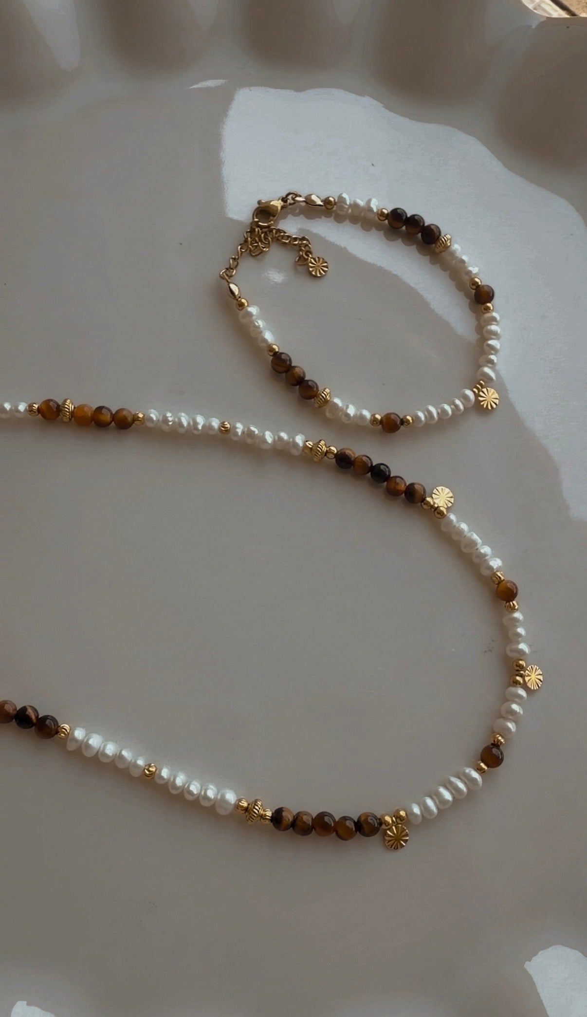 Handgefertigte Halskette und Armband aus Süsswasserperlen und Tigerauge mit vergoldeten Edelstahldetails auf Glasschale.