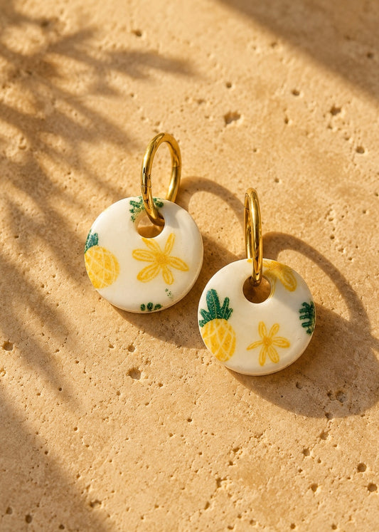Handgefertigte Polymer Clay Ohrringe mit Ananas Motiv an vergoldeter Edelstahl Creole auf Traversen im warmen Sonnenlicht. 