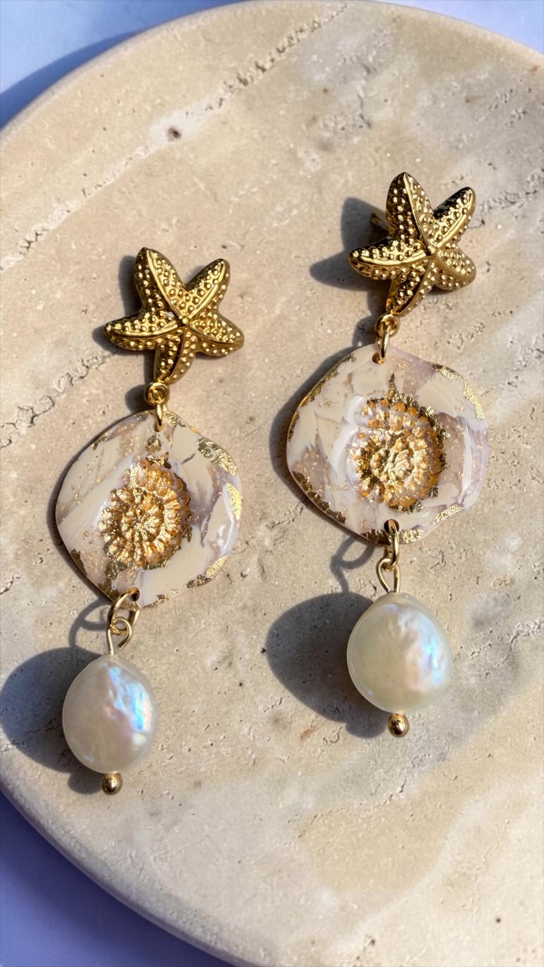 Ohrringe Nerea Pearl mit goldenem Seestern, marmorierter Polymer-Anhänger und Süsswasserperle, fotografiert auf  Travertinplatte