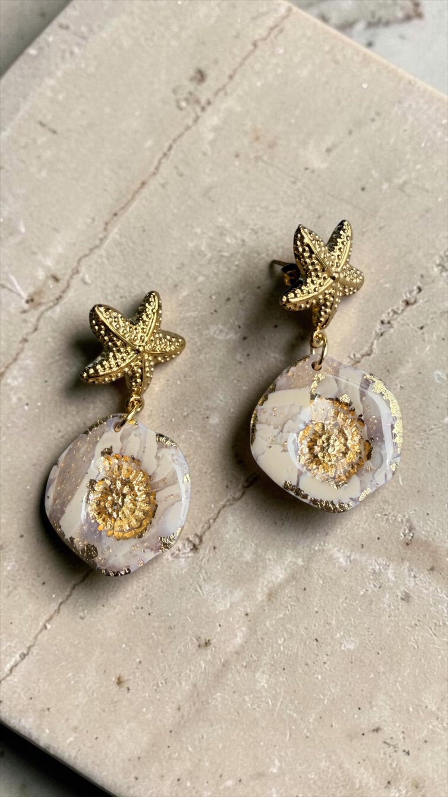 Ohrringe Nerea mit goldenem Seestern und marmorierter  Polymer Ton Anhänger mit Goldprint, fotografiert auf Travertinplatte